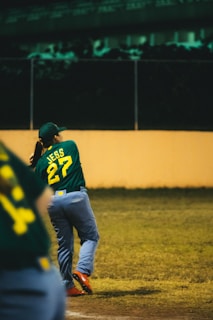 A baseball player in a green jersey with the number 27 swings a bat on a field. The player is wearing a cap and blue pants, and the field is surrounded by a fence with a dimly lit background.