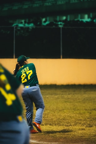 A baseball player in a green jersey with the number 27 swings a bat on a field. The player is wearing a cap and blue pants, and the field is surrounded by a fence with a dimly lit background.