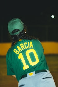 A person wearing a green baseball cap and jersey with the name 'Garcia' and the number '10'. The background is dimly lit, suggesting an outdoor setting at night, possibly a sports field.