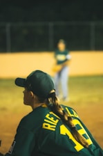 A person with a long braid is wearing a dark green baseball jersey with the number 10 and the name 'Rietveld' on it. They are seated or crouched on a baseball field, appearing to be focused on the game. Another player is visible in the blurred background wearing a similar uniform. The scene takes place at night under artificial lighting.