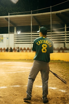A baseball player stands on a field, holding a bat. The player is wearing a green jersey with the name 'CASTAÑEDA' and number '8' on the back. The field is lined with a yellow barrier, and there are empty bleachers in the background under a partially covered roof. The scene appears to be at night, with artificial lighting illuminating the area.