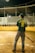 A baseball player stands on a field, holding a bat. The player is wearing a green jersey with the name 'CASTAÑEDA' and number '8' on the back. The field is lined with a yellow barrier, and there are empty bleachers in the background under a partially covered roof. The scene appears to be at night, with artificial lighting illuminating the area.