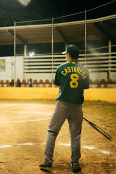 A baseball player stands on a field, holding a bat. The player is wearing a green jersey with the name 'CASTA&Ntilde;EDA' and number '8' on the back. The field is lined with a yellow barrier, and there are empty bleachers in the background under a partially covered roof. The scene appears to be at night, with artificial lighting illuminating the area.