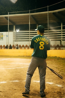 A baseball player stands on a field, holding a bat. The player is wearing a green jersey with the name 'CASTA&Ntilde;EDA' and number '8' on the back. The field is lined with a yellow barrier, and there are empty bleachers in the background under a partially covered roof. The scene appears to be at night, with artificial lighting illuminating the area.