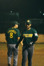 Two baseball players are on the field, wearing green jerseys and caps with logos. One player has the name 'Castaneda' and the number 8 on their back, while the other is facing forward with a serious expression. The background is dark, suggesting a nighttime game.