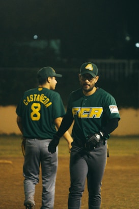 Two baseball players are on the field, wearing green jerseys and caps with logos. One player has the name 'Castaneda' and the number 8 on their back, while the other is facing forward with a serious expression. The background is dark, suggesting a nighttime game.