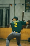 A baseball player wearing a green shirt with the number 2 and the word 'EAST' on the back is prepared to bat. The player is holding a bat in a ready position, standing in a batter's box. A chain-link fence is visible in the background, along with a yellow barrier and some colorful pennants.