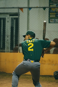 A baseball player wearing a green shirt with the number 2 and the word 'EAST' on the back is prepared to bat. The player is holding a bat in a ready position, standing in a batter's box. A chain-link fence is visible in the background, along with a yellow barrier and some colorful pennants.