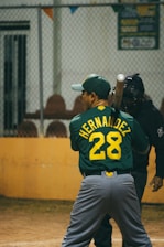 A baseball player wearing a green jersey with the name and number 'Hernandez 28' is preparing to bat. A catcher stands behind the player, dressed in black protective gear. They are on a baseball field, and the background has a metal fence and some seating.