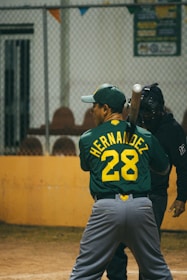 A baseball player wearing a green jersey with the name and number 'Hernandez 28' is preparing to bat. A catcher stands behind the player, dressed in black protective gear. They are on a baseball field, and the background has a metal fence and some seating.