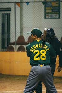 A baseball player wearing a green jersey with the name and number 'Hernandez 28' is preparing to bat. A catcher stands behind the player, dressed in black protective gear. They are on a baseball field, and the background has a metal fence and some seating.