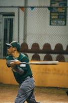 A baseball player in a green shirt and gray pants is captured mid-swing with a baseball bat. The setting appears to be a baseball field, with a chain-link fence and empty seats in the background. Banners or signs are visible above the fence.