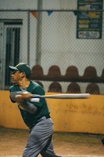 A baseball player in a green shirt and gray pants is captured mid-swing with a baseball bat. The setting appears to be a baseball field, with a chain-link fence and empty seats in the background. Banners or signs are visible above the fence.