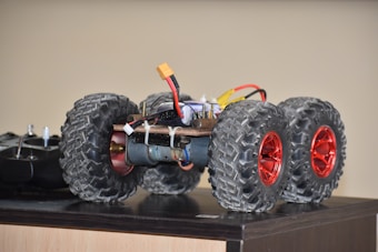 A small, six-wheeled vehicle or toy with large, rugged tires and red rims is placed on a tabletop. It has visible wiring and components, indicating it may be a battery-powered or remote-controlled car. The table surface is wooden, and there is a black remote control device next to the vehicle.