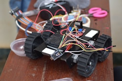 A small robotic vehicle is positioned on a wooden table, equipped with large black wheels and multiple colored wires connected to its top. Various electronic components are scattered around, including a pair of pink scissors, cables, and a small bowl.