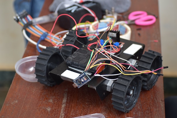 A small robotic vehicle is positioned on a wooden table, equipped with large black wheels and multiple colored wires connected to its top. Various electronic components are scattered around, including a pair of pink scissors, cables, and a small bowl.