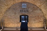 A brick archway with a sign that reads Vina Belje. In the center, there is a display case on a black-draped table. The interior has a rustic appearance with cylindrical metallic objects placed on either side of the archway.