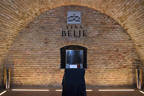 A brick archway with a sign that reads Vina Belje. In the center, there is a display case on a black-draped table. The interior has a rustic appearance with cylindrical metallic objects placed on either side of the archway.