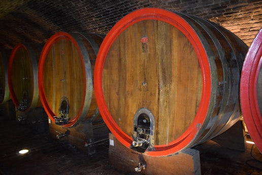 Large wooden barrels with red rims are lined up in a dimly lit wine cellar with a brick ceiling. Each barrel has a spigot at the bottom for dispensing liquid.
