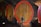Large wooden barrels with red rims are lined up in a dimly lit wine cellar with a brick ceiling. Each barrel has a spigot at the bottom for dispensing liquid.