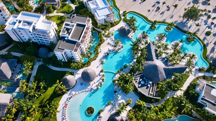 An aerial view of a luxurious resort featuring large swimming pools with curved shapes surrounded by lush green palm trees. Several modern white buildings with multiple stories are visible, accompanied by thatched-roof structures and sun lounges lining the poolsides. The landscape exhibits a mix of organized pathways and tropical vegetation.