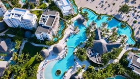 An aerial view of a luxurious resort featuring large swimming pools with curved shapes surrounded by lush green palm trees. Several modern white buildings with multiple stories are visible, accompanied by thatched-roof structures and sun lounges lining the poolsides. The landscape exhibits a mix of organized pathways and tropical vegetation.