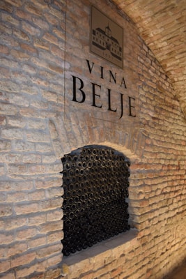 A textured brick wall with a large recessed area filled with stacked wine bottles. Above the recess, a sign reads 'Vina Belje,' accompanied by an emblem or logo, possibly depicting a building and the year 1697.