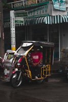 A tricycle with a yellow frame and decorative elements is parked on a wet street. A person wearing a bright red shirt with text is seated inside the vehicle. The background shows a storefront with green and white signage and a green awning.