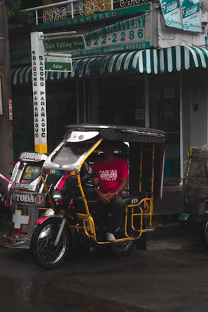 A tricycle with a yellow frame and decorative elements is parked on a wet street. A person wearing a bright red shirt with text is seated inside the vehicle. The background shows a storefront with green and white signage and a green awning.