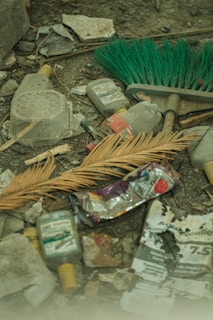A cluttered scene featuring discarded plastic bottles, a crushed metal can, a broom with green bristles, a dried palm leaf, and torn paper and debris scattered on the ground. The setting appears to be outdoors or in an abandoned area.