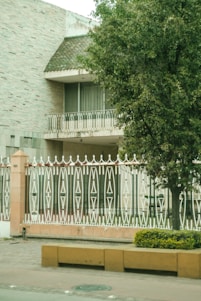 A house with a textured brick exterior and a sloped roof featuring small green tiles. A balcony with ornate white metal railings is visible, along with a large tree providing shade. In the foreground, a decorative fence with intricate geometric patterns stands behind a low wall. A yellow planter with shrubs adorns the pavement.