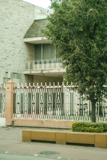 A house with a textured brick exterior and a sloped roof featuring small green tiles. A balcony with ornate white metal railings is visible, along with a large tree providing shade. In the foreground, a decorative fence with intricate geometric patterns stands behind a low wall. A yellow planter with shrubs adorns the pavement.