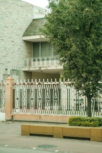 A house with a textured brick exterior and a sloped roof featuring small green tiles. A balcony with ornate white metal railings is visible, along with a large tree providing shade. In the foreground, a decorative fence with intricate geometric patterns stands behind a low wall. A yellow planter with shrubs adorns the pavement.