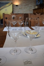 Two wine glasses placed on a table are set in an arranged manner. There's a soft light illuminating the background with a brick wall, creating a cozy ambiance. The table setting includes tasting mats and plates, suggesting a wine tasting or similar event.
