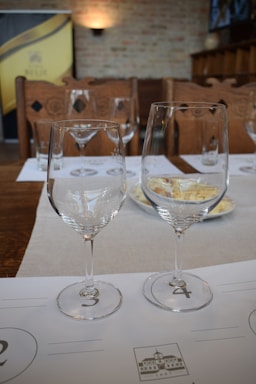Two wine glasses placed on a table are set in an arranged manner. There's a soft light illuminating the background with a brick wall, creating a cozy ambiance. The table setting includes tasting mats and plates, suggesting a wine tasting or similar event.