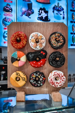 A variety of nine colorful and creatively decorated donuts are displayed on a wooden board, each with distinct toppings and designs, set against a backdrop of illustrated menu items on a screen.