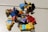 Several LEGO minifigures are scattered on a light-colored surface. The figures display various designs, including one dressed in a red outfit with black markings and another with a purple and black cape. A variety of accessories, such as helmets and colored items, are visible among the figures.