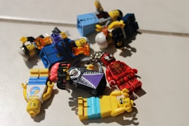 Several LEGO minifigures are scattered on a light-colored surface. The figures display various designs, including one dressed in a red outfit with black markings and another with a purple and black cape. A variety of accessories, such as helmets and colored items, are visible among the figures.