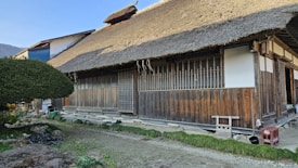 A traditional wooden house with a thatched roof is surrounded by a serene natural setting. The building features wooden sliding doors, a white plastered section, and a small entrance area. A neatly trimmed tree is positioned nearby along with other greenery.