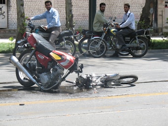 A motorcycle is lying on its side in the street, possibly indicating an accident. Three men on motorcycles are nearby, observing the scene. The road shows signs of damage or fluid leakage, and there are trees and a brick wall in the background.