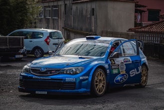 A blue rally car with numerous sponsor logos is parked on a rough, paved surface in an outdoor setting, alongside another vehicle. The car's doors are open, and it has a sporty, aggressive design typical of performance vehicles meant for racing. The background includes a building with a rustic appearance and a fence.