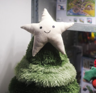 A soft, plush Christmas tree topped with a smiling star is displayed, likely inside a store. The tree is made from a deep green, fluffy material with multiple layers. Behind it, there are shelves with various other items, creating a casual, festive atmosphere.