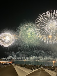 Vibrant fireworks burst in various patterns against a dark night sky, illuminating a waterfront scene. Reflections of colorful fireworks can be seen on the water. In the foreground, beach chairs and umbrellas are visible, suggesting a coastal location.