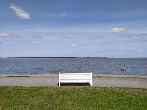 A white bench is placed on a grassy area facing a calm body of water, with a clear blue sky dotted with a few clouds overhead. The horizon appears distant, creating a sense of openness and tranquility.