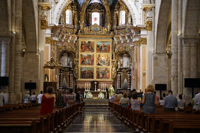 A grand interior of a cathedral showcasing intricate architecture with ornate gold detailing and large, colorful religious paintings. Several people are gathered in the pews, with a priest standing at the altar.