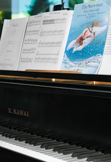 A close-up view of a black K. Kawai piano with sheet music on the music stand. The sheets include musical notes and a cover page for 'The Snowman Easy Piano Suite' featuring an illustration of a snowman and a child flying in the sky. The background displays a blurred green tree seen through a window.