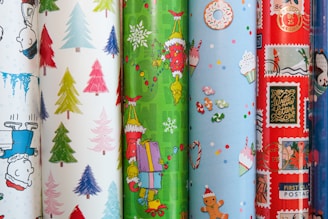 A collection of colorful, festive wrapping papers featuring various holiday-themed designs, such as cartoon characters, Christmas trees, snowflakes, cupcakes, candies, and postal motifs. The designs vary from cute and playful to vintage and classic, with diverse textures and patterns.