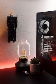A modern desk setup includes a glowing lamp with a vintage bulb design, a small potted succulent plant, and a toy car model on a dark surface. A computer case with visible fans and hardware is on the right, featuring RGB lighting. On the wall, a decorative piece resembles a bat with a flame effect.