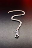 A silver chain with an intricate design lies on a dark surface. Attached to the chain is a metal pendant shaped like a zipper pull with engraved lettering. Next to the pendant, there is a small circular charm with an embossed logo.