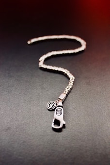 A silver chain with an intricate design lies on a dark surface. Attached to the chain is a metal pendant shaped like a zipper pull with engraved lettering. Next to the pendant, there is a small circular charm with an embossed logo.